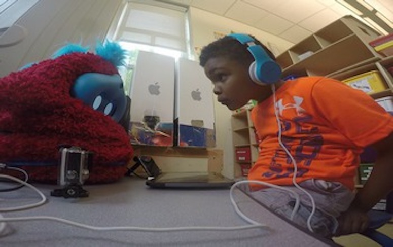A boy participates in a study with a fluffy red Tega robot. He has blue headphones on and is staring very intently at the robot.