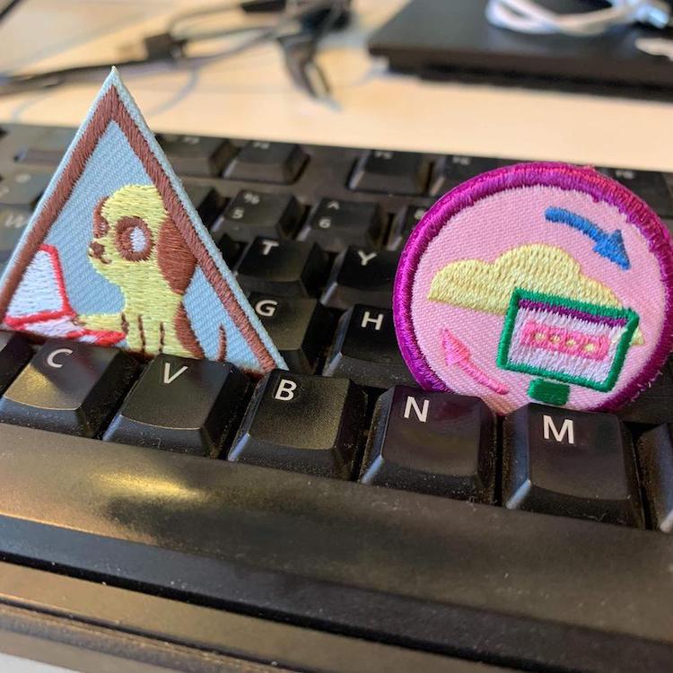 Two Girl Scout badges resting on a keyboard. One is triangular and shows a dog on a laptop, while the other is circular and shows a cycle between a monitor and a cloud.