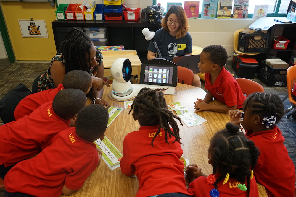 Group of children in red uniform shirts and a teacher gather around a Jibo robot and a tablet while researcher looks on