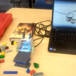 A smiling robot made out of Legos is plugged into a laptop
