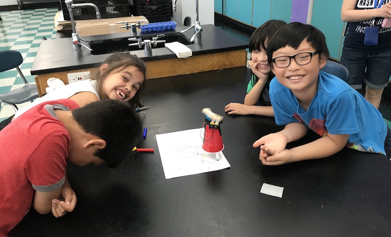 Four children laugh at a robot made out of a red solo cup and batteries.