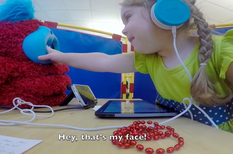 A smiling girl wearing blue headphones reaches out to squeeze the face of a fluffy red Tega robot. The robot is replying, 'Hey, that's my face!'