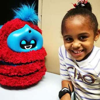 Girl smiling alongside red fluffy Tega robot.