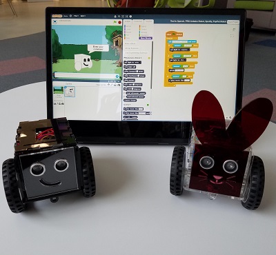 Two cute robots with smiley faces and eyes sit in front of a tablet with Scratch blocks loaded onto it.